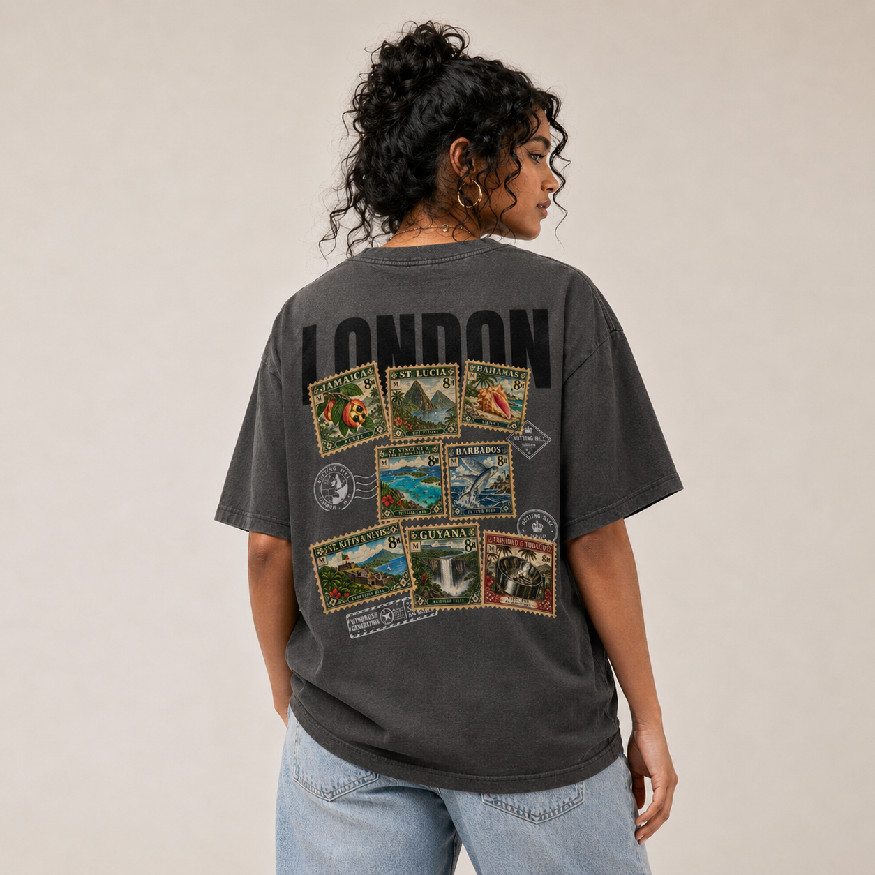 A woman seen from behind, hair pinned up, wearing the washed charcoal Maknesan tee with light denim. The back print reads LONDON in heavy black type above a collage of Caribbean postage stamps — Jamaica, St. Lucia, Bahamas, St. Vincent, Barbados, St. Kitts & Nevis, Guyana, Trinidad & Tobago — franked with round Notting Hill postmarks.